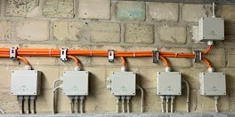 Pomarańczowe przewody w metalowych uchwytach prowadzą do białych puszek na nierównej, kremowej ścianie z cegieł. Widoczne kable wychodzące z puszek.