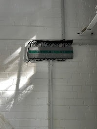 Szara skrzynka elektryczna z zielonymi elementami, zamontowana na białej, ceglanej ścianie. Widoczne kable i rury instalacyjne. Oświetlenie boczne tworzy cienie.