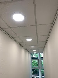 Równomiernie rozmieszczone, okrągłe lampy sufitowe LED w korytarzu z oknem. Białe ściany i sufit podwieszany z widoczną kratownicą. Ujęcie z dołu, perspektywa zbieżna.