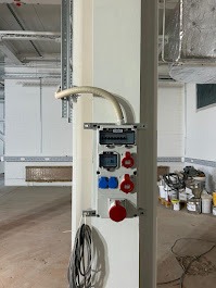 Biała skrzynka elektryczna z gniazdami (czerwone i niebieskie) i bezpiecznikami, zamontowana na kolumnie w surowym wnętrzu. Widoczne kable i elementy instalacji.