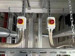 Dwa wyłączniki bezpieczeństwa z czerwonymi pokrętłami zamontowane na metalowej konstrukcji pod sufitem, z widocznymi kablami i rurami wentylacyjnymi.