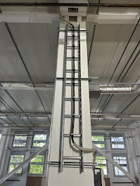 Wysoka kolumna z zamocowanymi drabinkami kablowymi i rurami osłonowymi w jasnym, industrialnym wnętrzu. Widoczne okna i elementy wentylacji pod sufitem.