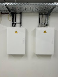 Dwie białe skrzynki elektryczne z żółtym symbolem ostrzegawczym, zamontowane na ścianie pod metalową korytką kablową z przewodami.