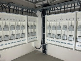 Wnęka z zamontowanymi, szarymi licznikami elektrycznymi w białej obudowie. Kable prowadzone w korytkach pod sufitem. Widok z dołu, oświetlenie sufitowe.