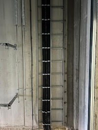 Kable w czarnej karbowanej osłonie ułożone w metalowej drabince kablowej, zamocowanej do surowej ściany. Widoczne uchwyty i inne przewody. Ujęcie z dołu.