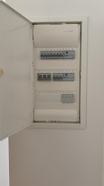 Otwarta, biała skrzynka elektryczna z bezpiecznikami na białej ścianie. Widoczne zabezpieczenia i listwy. Ujęcie z boku, lekko od dołu.