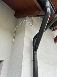 Biała, zewnętrzna instalacja elektryczna poprowadzona po elewacji budynku, obok czarnej rynny. Widok z dołu, lekko zniekształcony perspektywą.