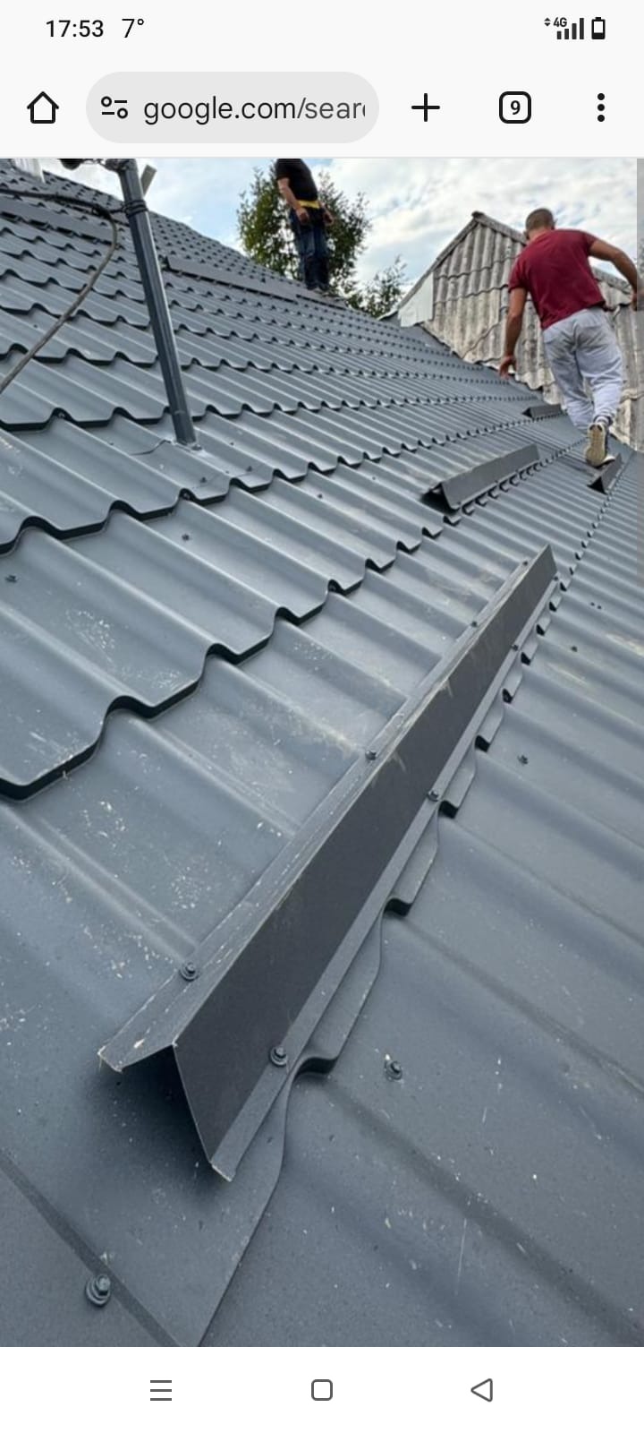 Szary dach z blachy trapezowej z zamontowanymi obróbkami blacharskimi. Dwóch dekarzy na dachu podczas pracy. Widok z dołu na ukończony fragment dachu.