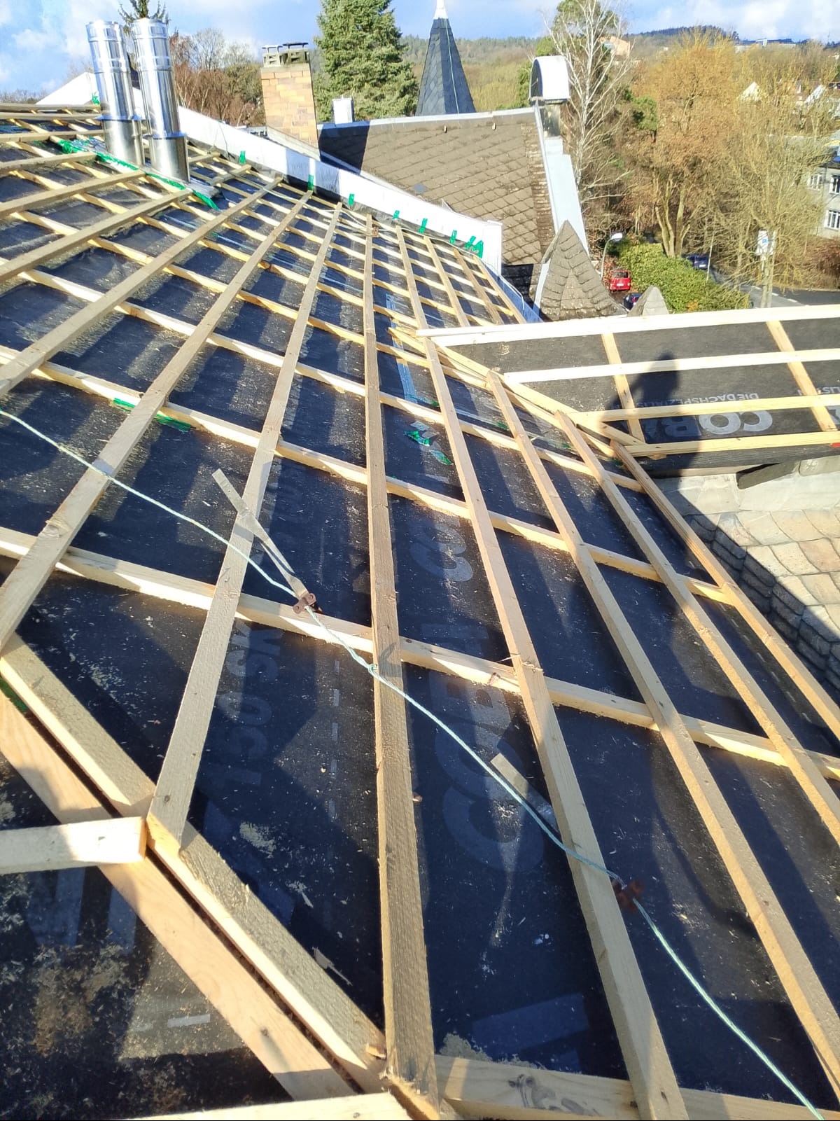 Konstrukcja dachu z drewnianymi łatami i membraną dachową w trakcie montażu. Widoczne kominy i fragmenty budynków w tle. Ujęcie z góry, w słoneczny dzień.