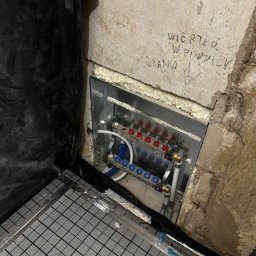 Jakub Szczypkowski - Rozdzielacz ogrzewania podłogowego w metalowej obudowie, zamontowany w surowej wnęce ściany, z widoczną izolacją i folią pod wylewkę. Napis na ścianie.