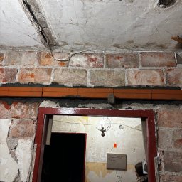 Jakub Szczypkowski - Ceglana ściana z widocznymi przewodami i rurami, surowy strop z instalacją elektryczną, czerwona futryna drzwi. Wnętrze w trakcie remontu, widoczne ślady po instalacjach.