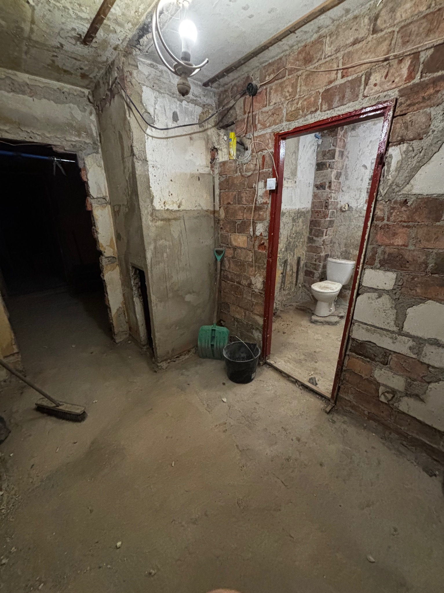Surowe wnętrze łazienki w trakcie remontu: odsłonięta cegła, stara toaleta, widoczne narzędzia i wiadro. Etap przygotowawczy do dalszych prac wykończeniowych.