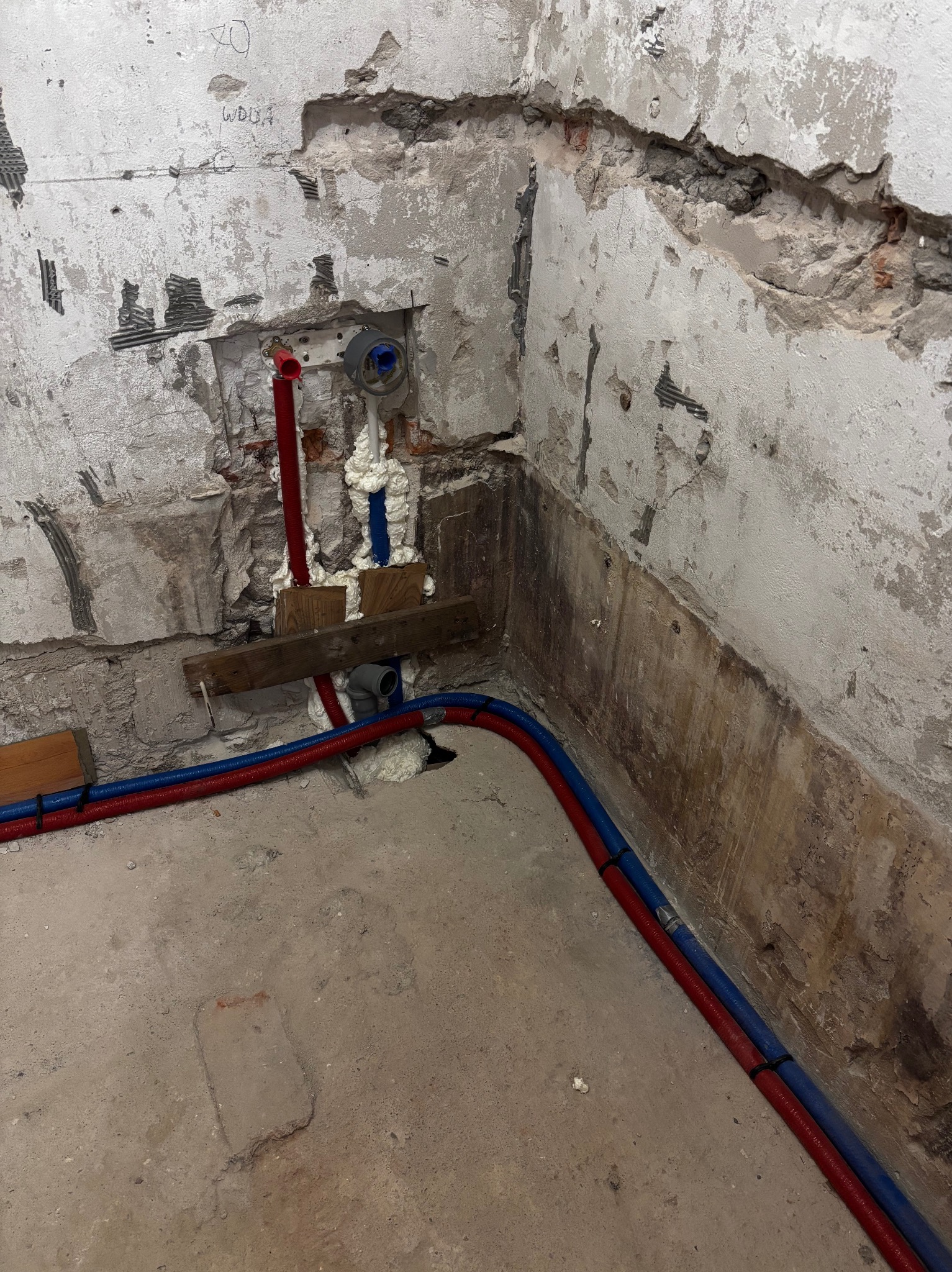 Instalacja wodno-kanalizacyjna w trakcie remontu łazienki: rury czerwone i niebieskie, pianka montażowa, surowe ściany z widocznymi śladami po płytkach.