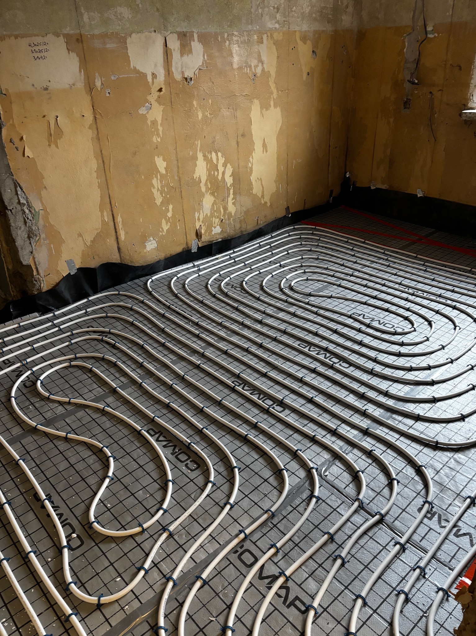Białe rury ogrzewania podłogowego ułożone spiralnie na czarnej macie z siatką, widoczne fragmenty ścian w trakcie remontu. Instalacja przed wylaniem posadzki.