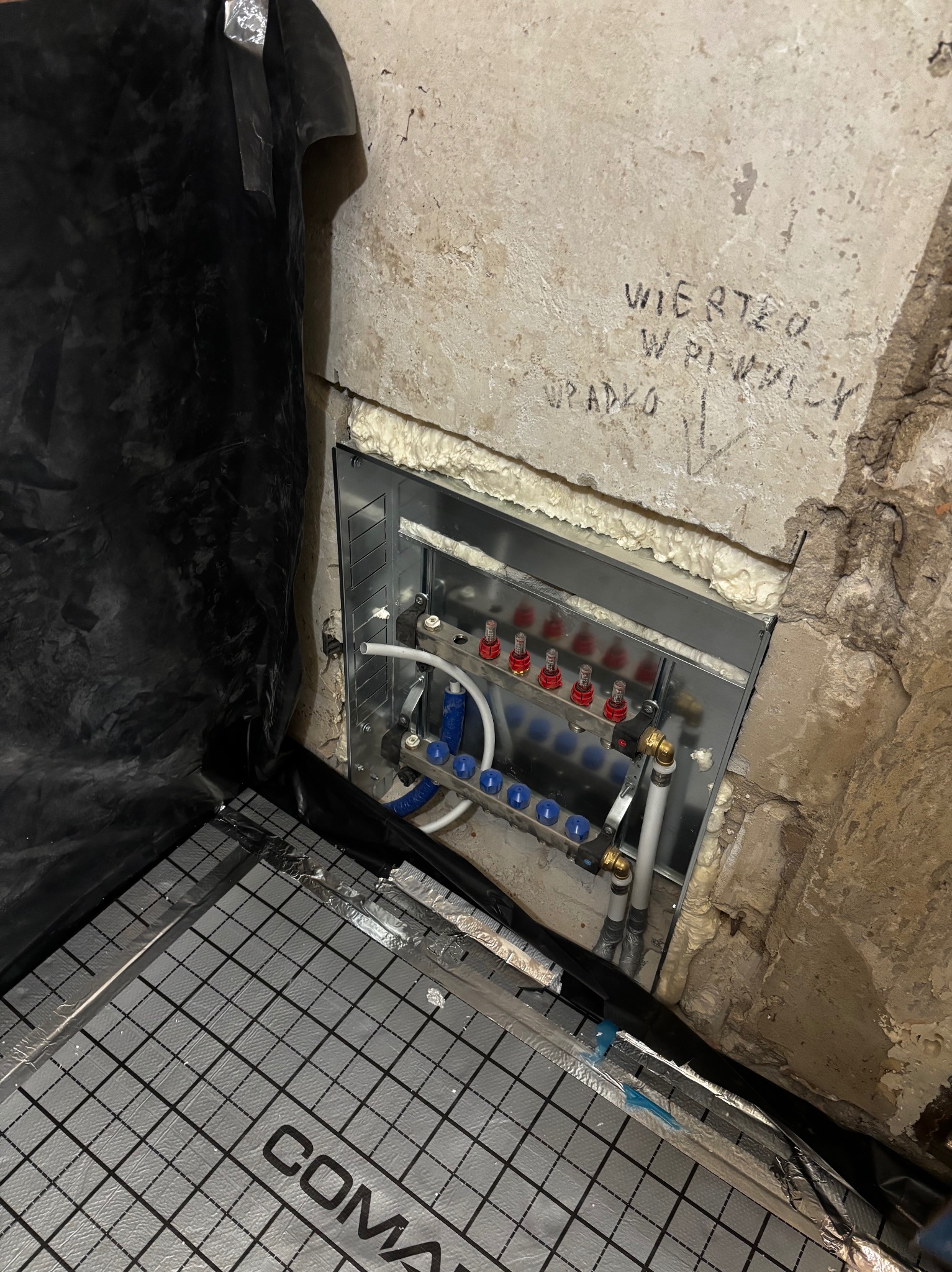 Rozdzielacz ogrzewania podłogowego w metalowej obudowie, zamontowany w surowej wnęce ściany, z widoczną izolacją i folią pod wylewkę. Napis na ścianie.