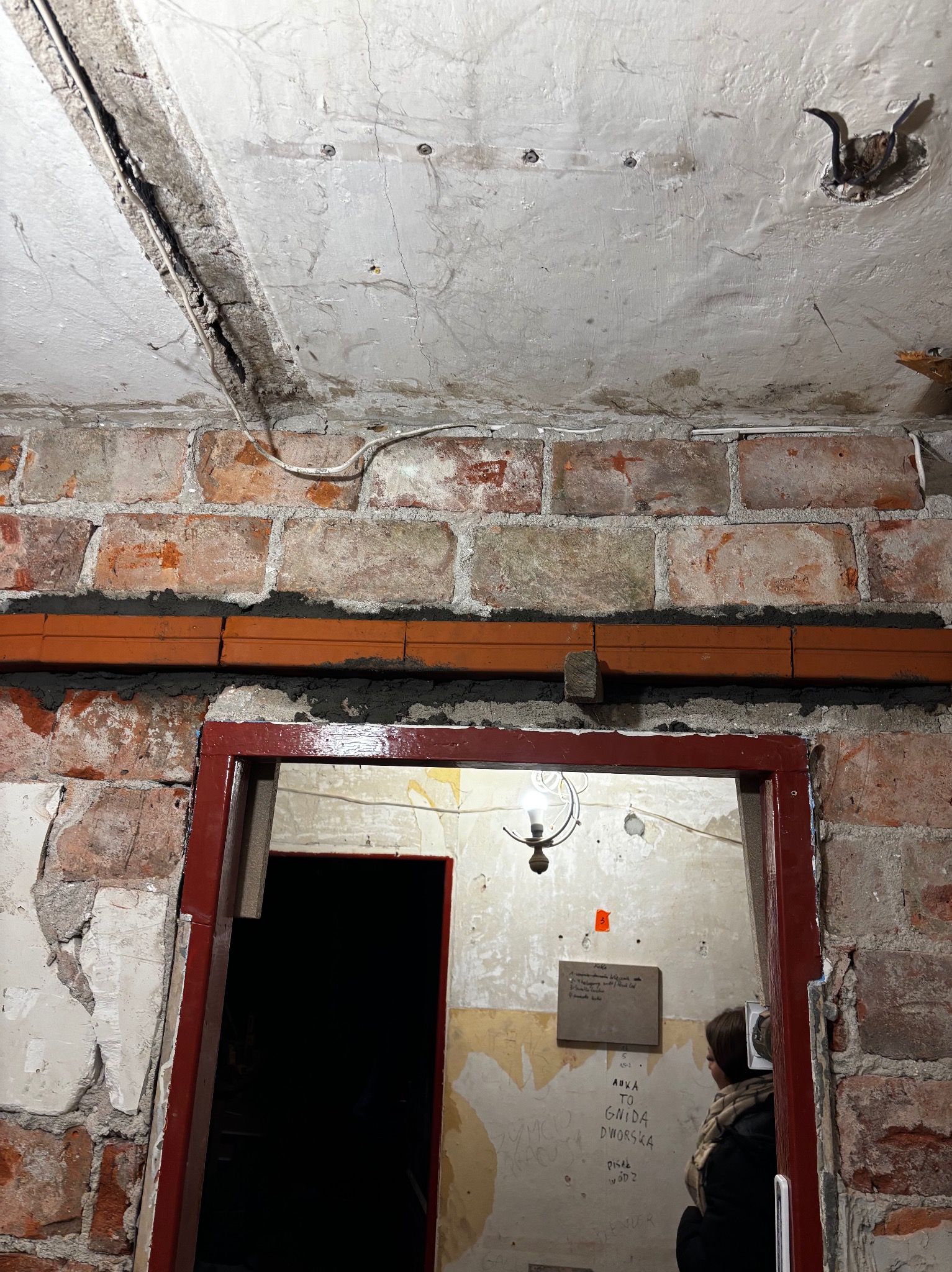 Ceglana ściana z widocznymi przewodami i rurami, surowy strop z instalacją elektryczną, czerwona futryna drzwi. Wnętrze w trakcie remontu, widoczne ślady po instalacjach.