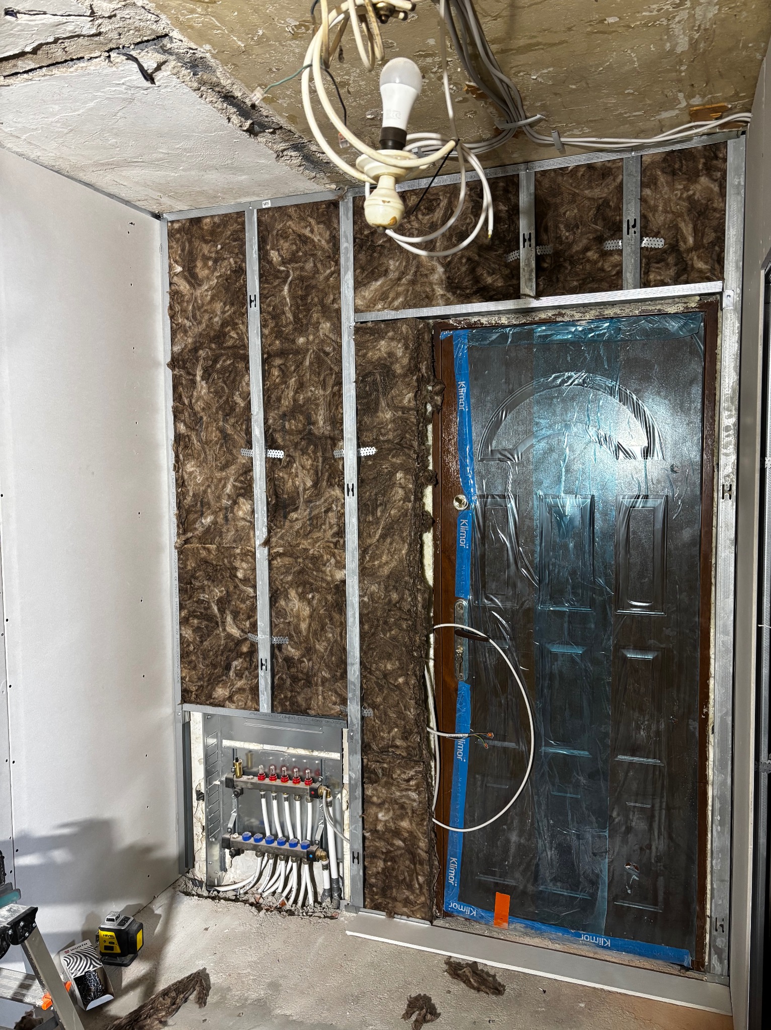 Ocieplanie ściany wełną mineralną, widoczna instalacja ogrzewania podłogowego i zabezpieczone folią drzwi wejściowe w trakcie prac wykończeniowych.