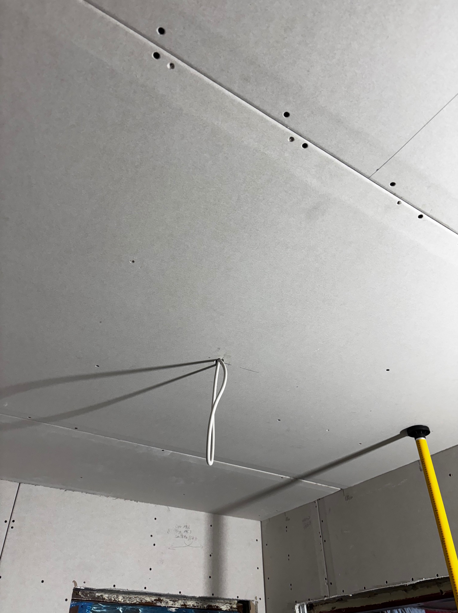 Zamontowane płyty gipsowo-kartonowe na suficie, widoczne ślady po wkrętach, kabel zwisający z otworu, żółta podpórka teleskopowa. Prace wykończeniowe.