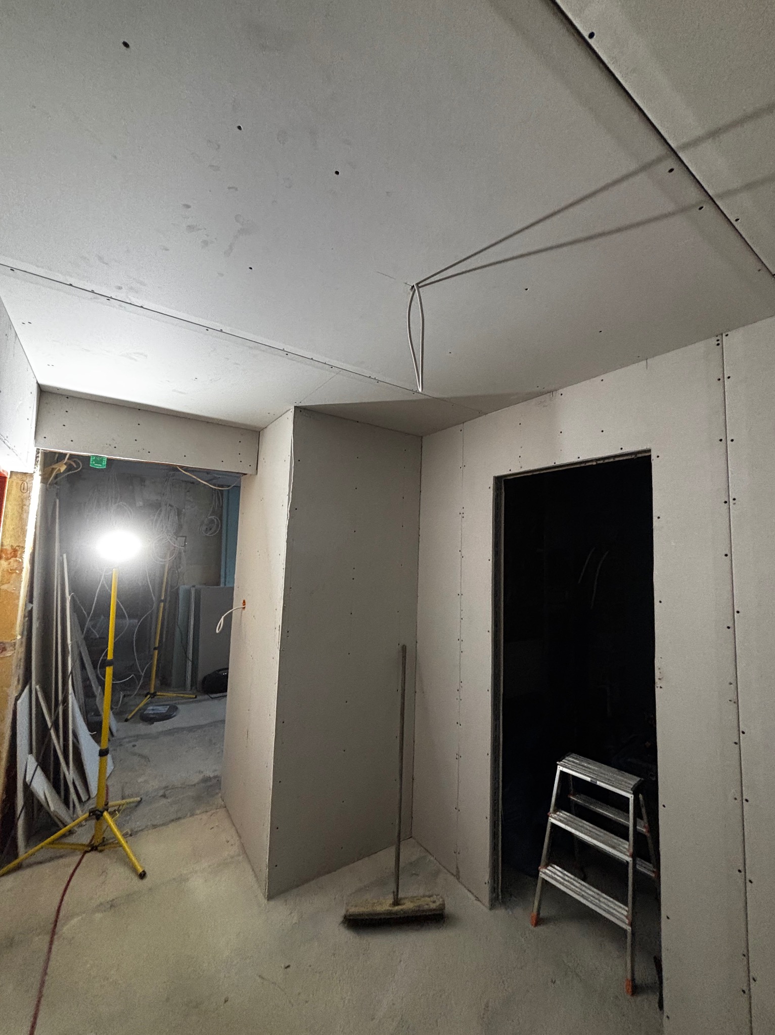Wnętrze w trakcie montażu płyt gipsowo-kartonowych, widoczna drabina, lampa budowlana i szczotka. Surowy stan wykończenia, przewody elektryczne.