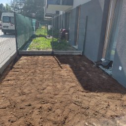 Aranżacja ogrodów - Przygotowany teren pod trawnik z widoczną siatką wzmacniającą, obok budynku z szarą elewacją i metalowym ogrodzeniem. W tle zieleń i zaparkowany bus.