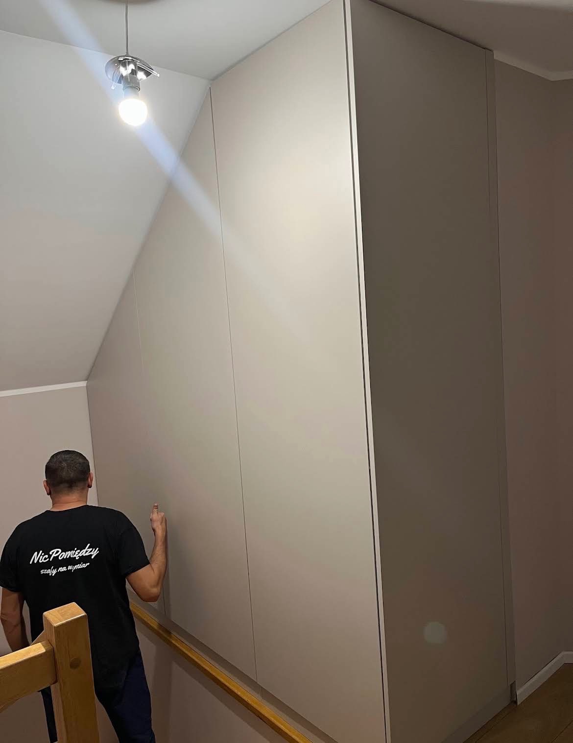 Jasna szafa wnękowa na poddaszu ze skośnym sufitem, dopasowana do przestrzeni. Mężczyzna w czarnej koszulce z napisem wskazuje na front szafy.