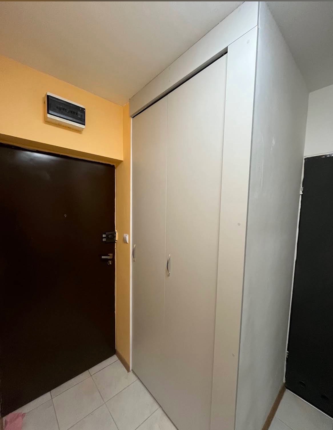 Biała szafa przesuwna z trzema drzwiami, wkomponowana we wnękę przy wejściu do mieszkania. Widoczna brązowa ściana i drzwi wejściowe.