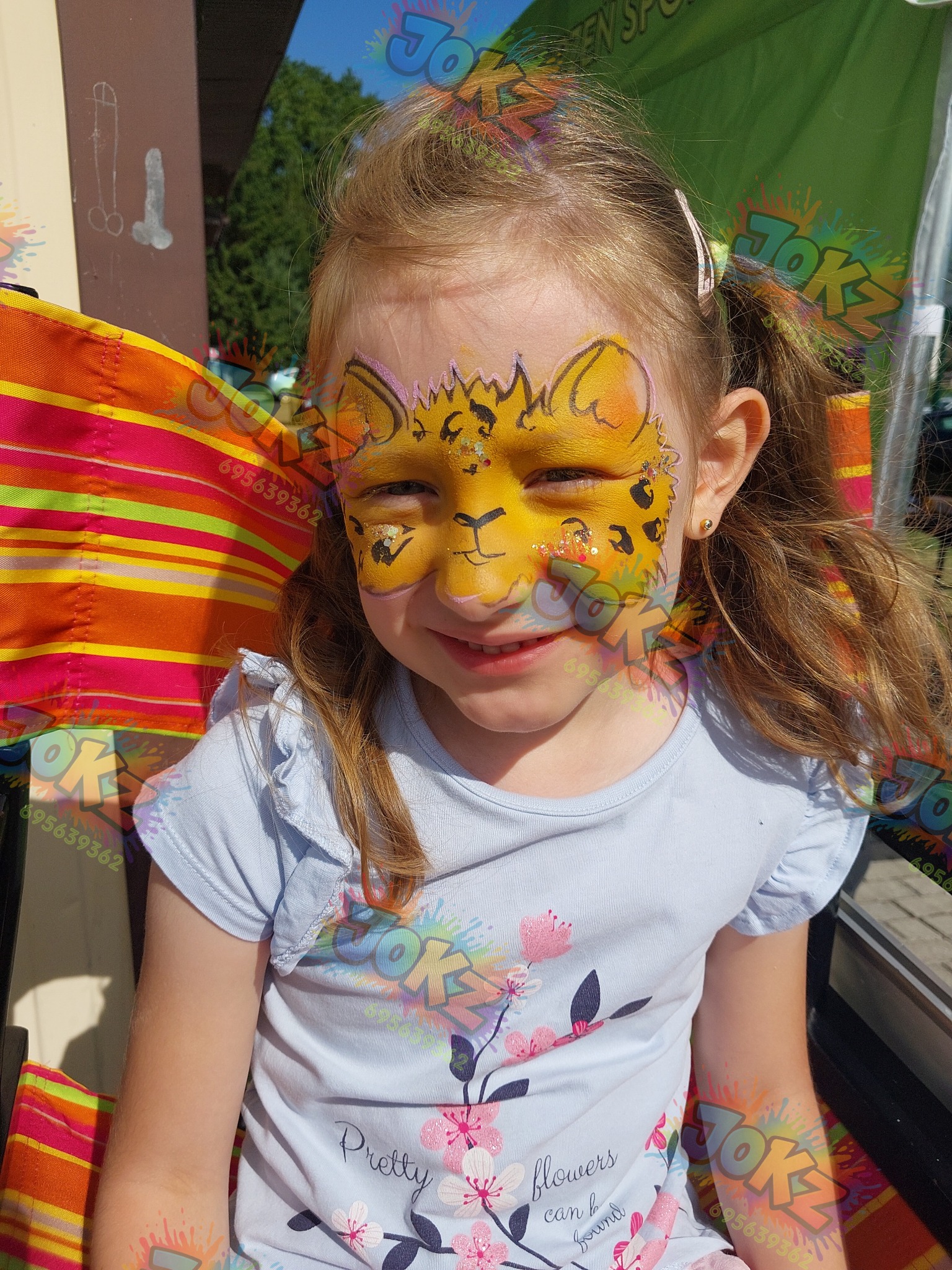 Uśmiechnięta dziewczynka z pomalowaną twarzą w motywy geparda, siedząca na krześle plażowym w słoneczny dzień. Widoczne brokatowe akcenty na twarzy.