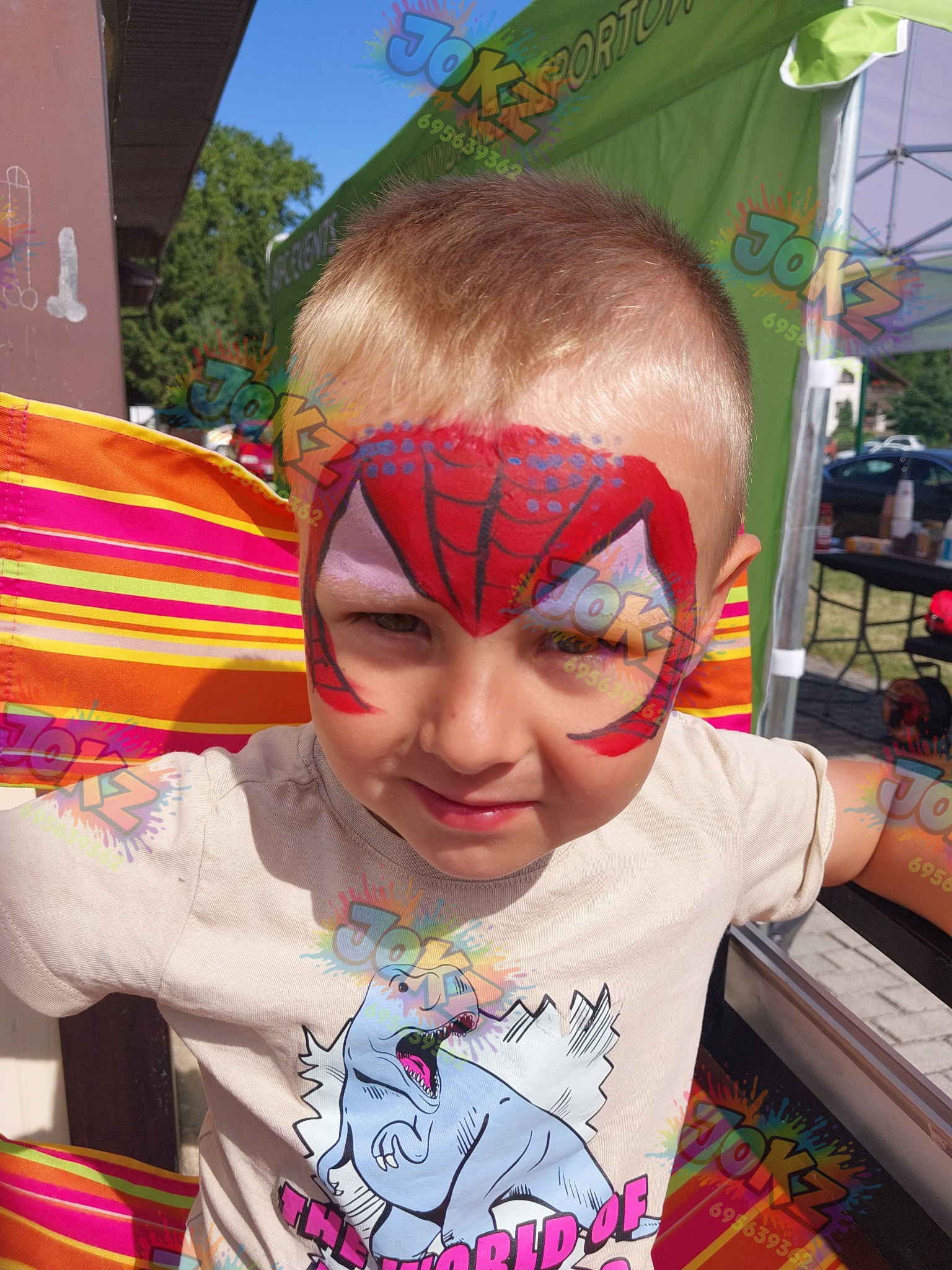 Uśmiechnięty chłopiec z pomalowaną twarzą w motywie Spider-Mana, siedzący na kolorowym krześle, w tle zielony namiot i drzewa. Wyraźne detale malunku.