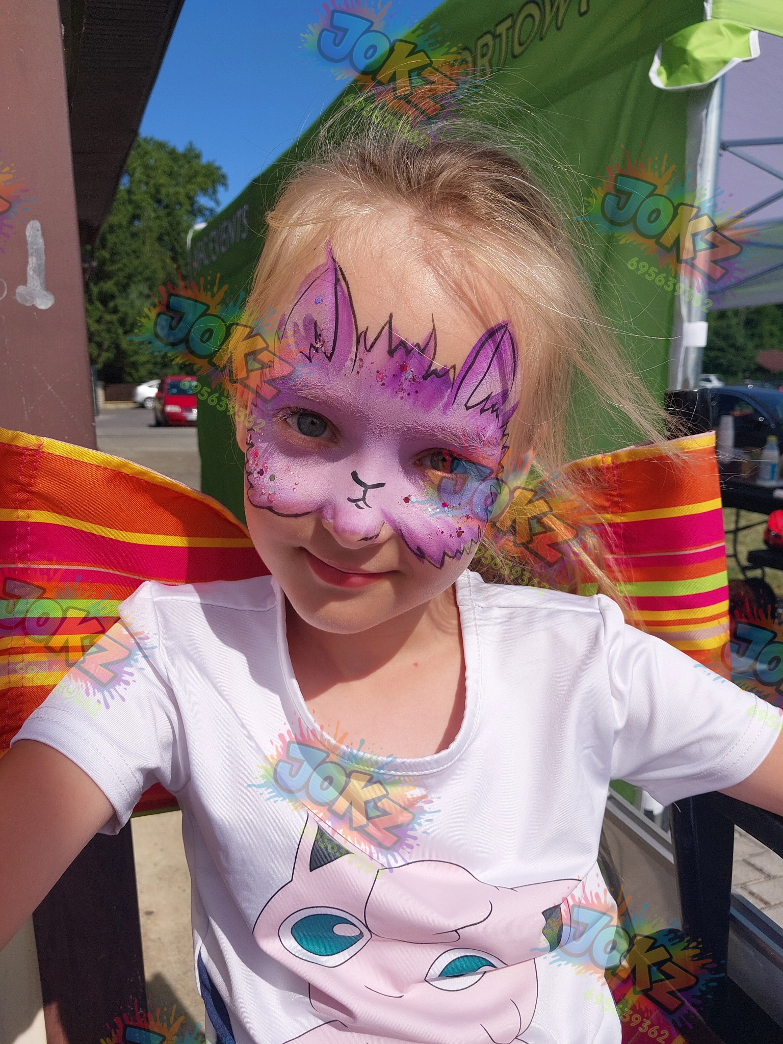 Uśmiechnięta dziewczynka z fioletowym makijażem twarzy w kształcie kota, siedząca na krześle z kolorowym oparciem, w koszulce z motywem Jigglypuff.