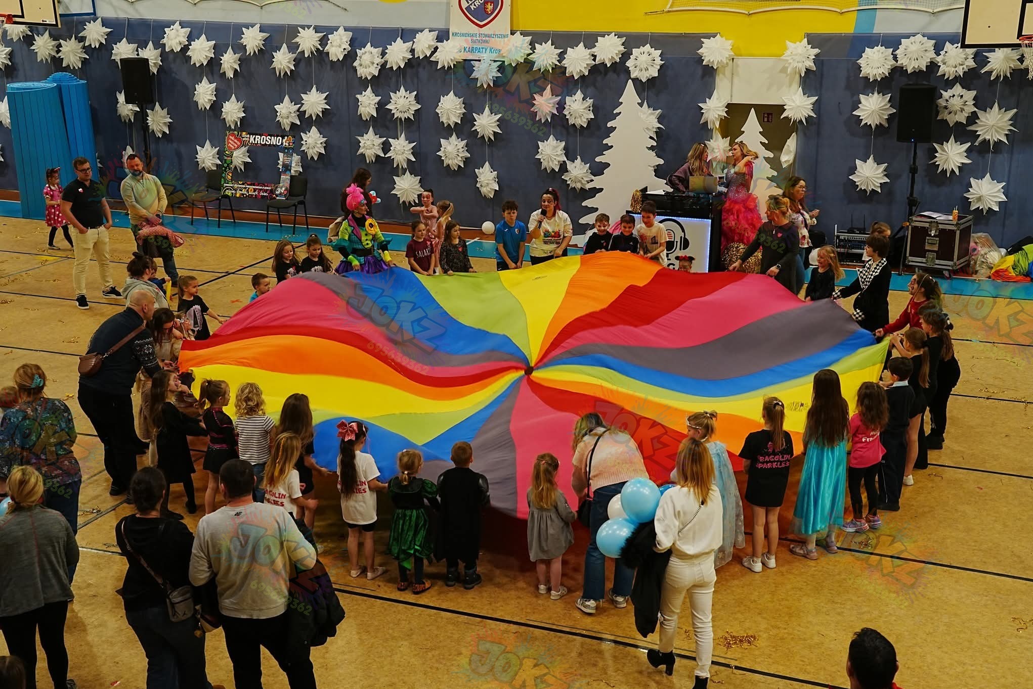 Animator z dziećmi bawi się kolorową chustą animacyjną podczas imprezy w sali gimnastycznej, w tle dekoracje i DJ. Zabawa z chustą animacyjną.