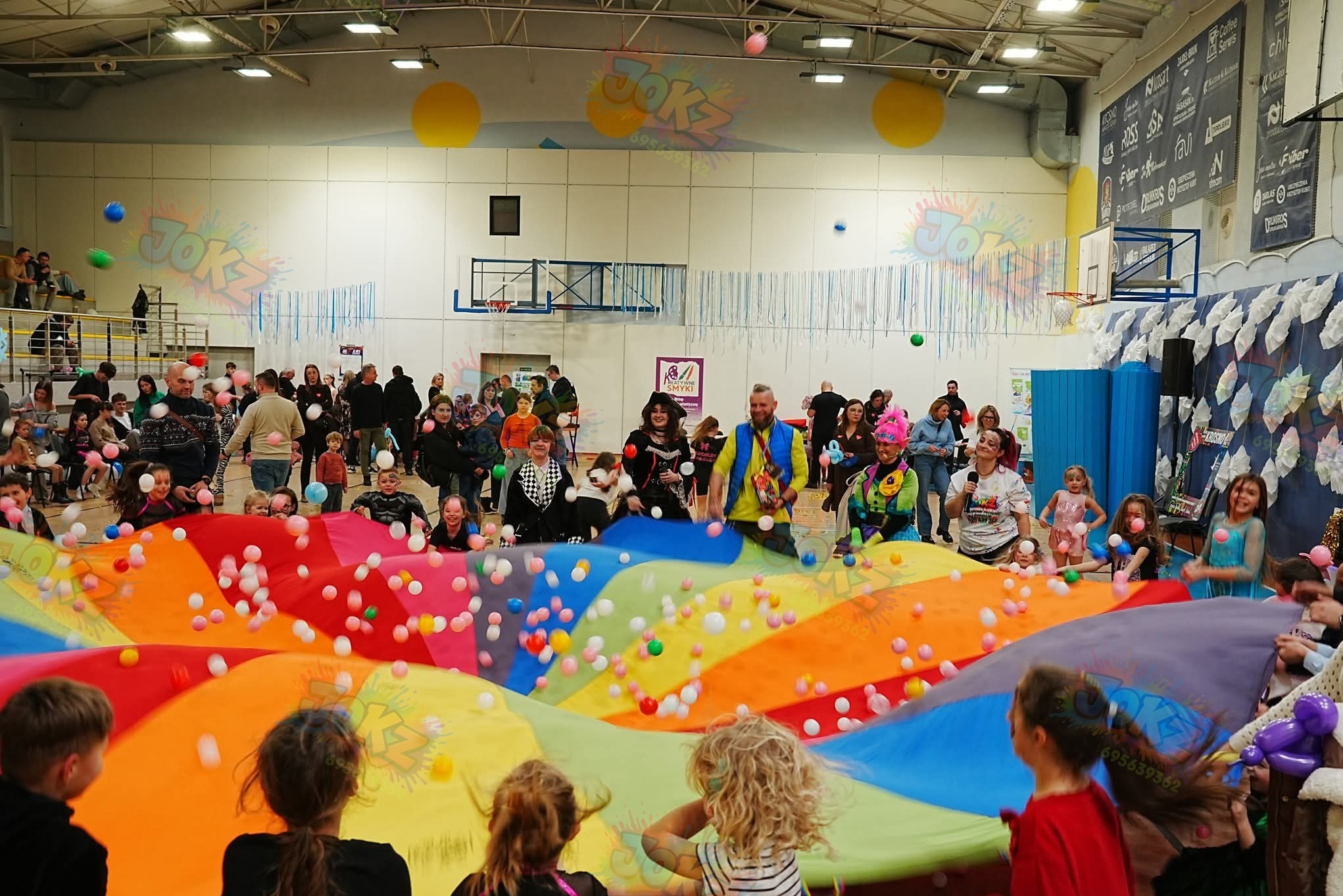 Animatorzy w akcji z dziećmi, używając kolorowej chusty animacyjnej i piłeczek w sali gimnastycznej. Zabawa z elementami edukacyjnymi i integracyjnymi.