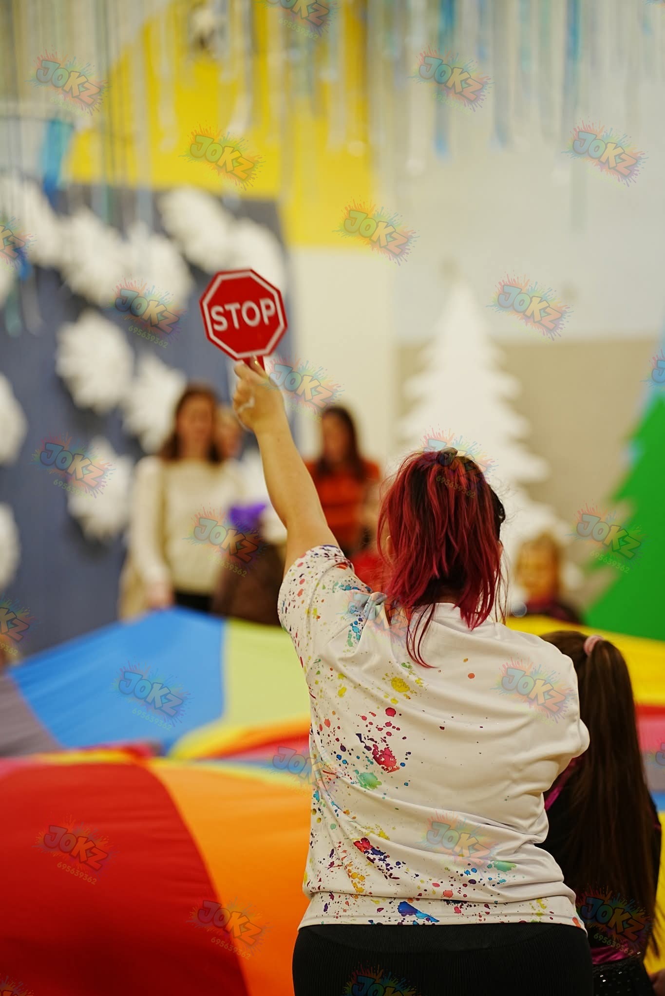 Animator z czerwoną czupryną trzyma znak STOP podczas zabawy z dziećmi na kolorowej macie w sali zabaw. W tle dekoracje i inne osoby.