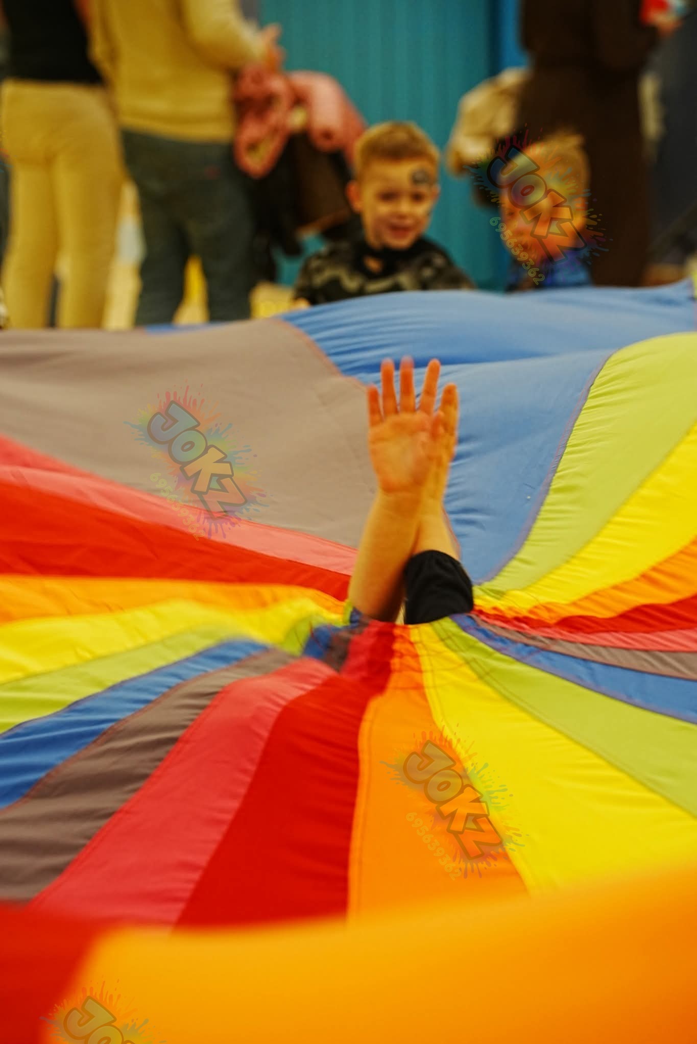 Dzieci bawią się pod kolorową chustą animacyjną, unosząc ręce do góry. Widoczne twarze uśmiechniętych chłopców z pomalowanymi buziami. Dynamiczne ujęcie z perspektywy uczestnika zabawy.
