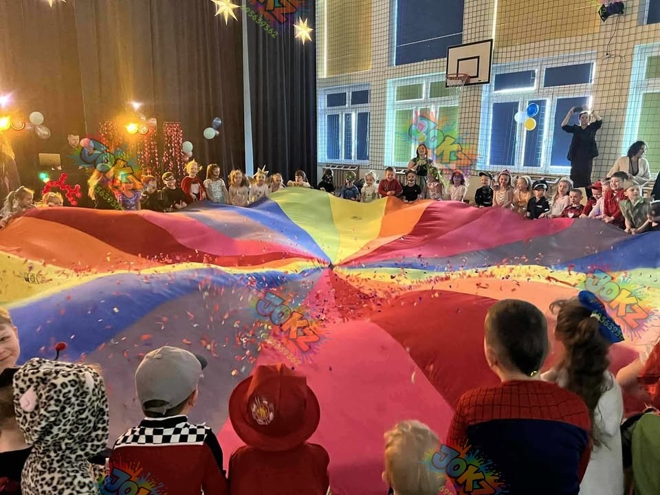 Animatorzy i dzieci bawią się kolorową chustą animacyjną w sali gimnastycznej podczas imprezy. Konfetti w powietrzu, uśmiechy, kostiumy.