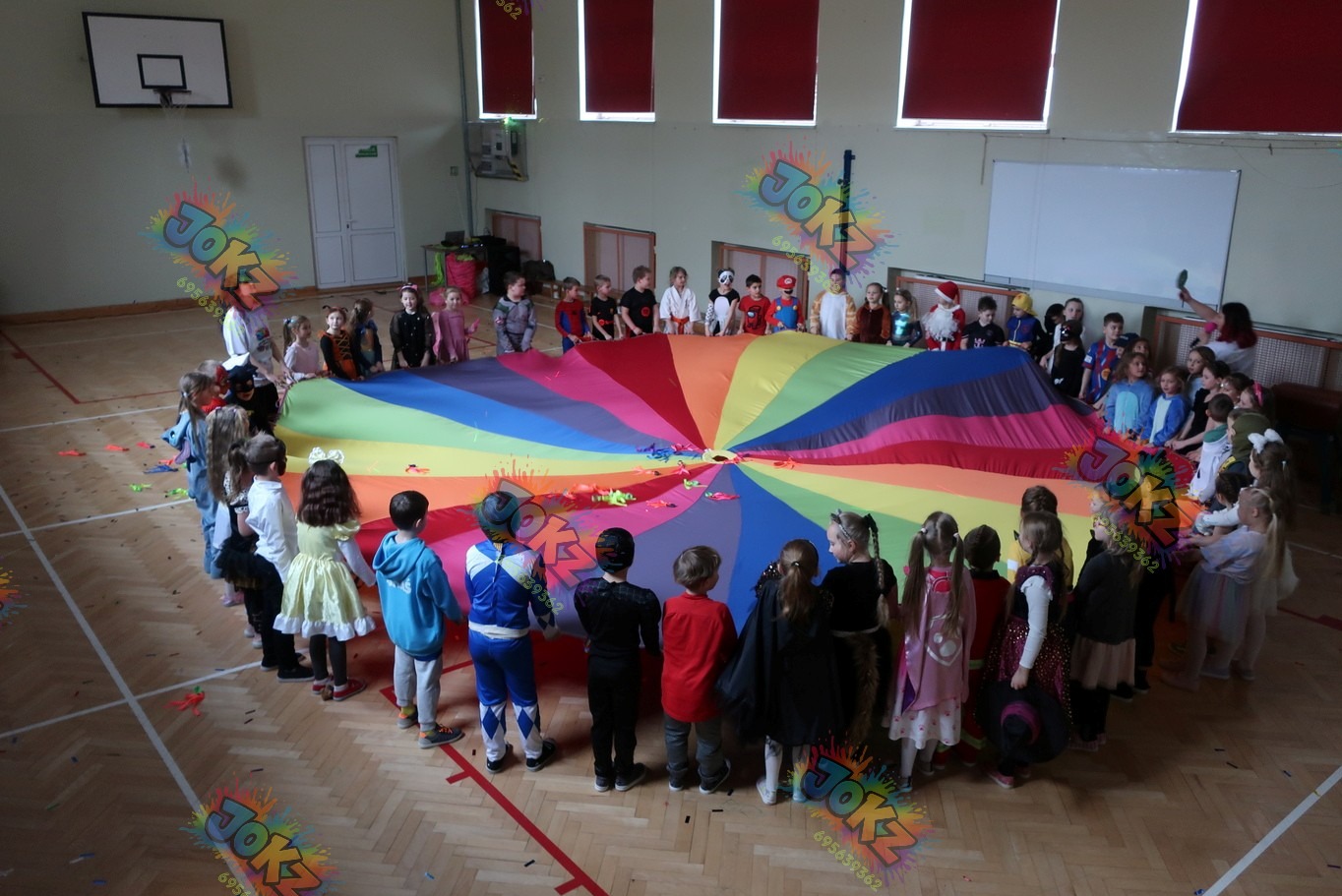 Grupka dzieci w kostiumach bawi się kolorową chustą animacyjną w sali gimnastycznej. Widoczni animatorzy i elementy dekoracji. Zabawa z chustą animacyjną.