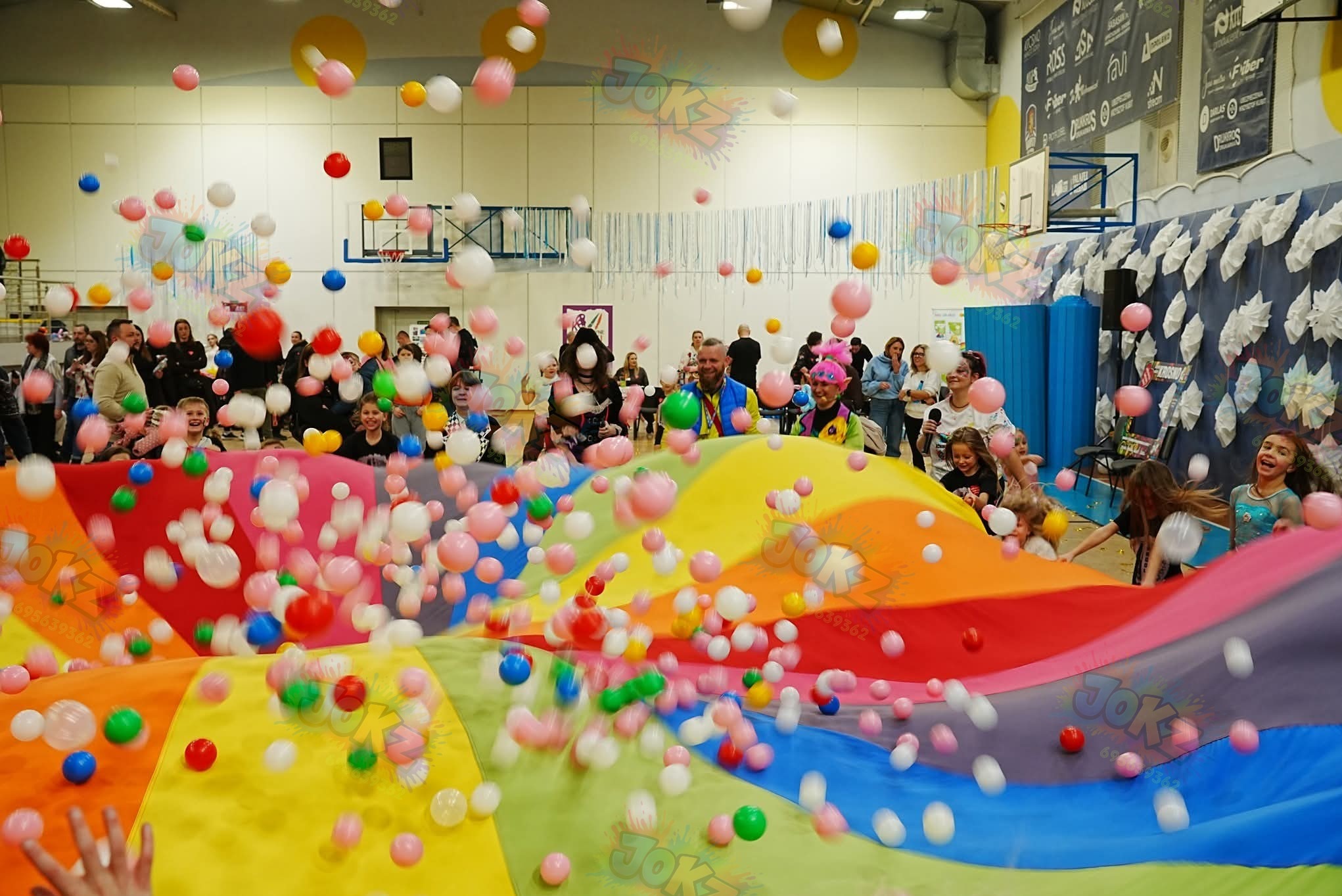 Radosna zabawa z kolorową chustą animacyjną i deszczem piłeczek w sali gimnastycznej. Dzieci i animatorzy w akcji, dynamiczne ujęcie z perspektywy uczestnika.