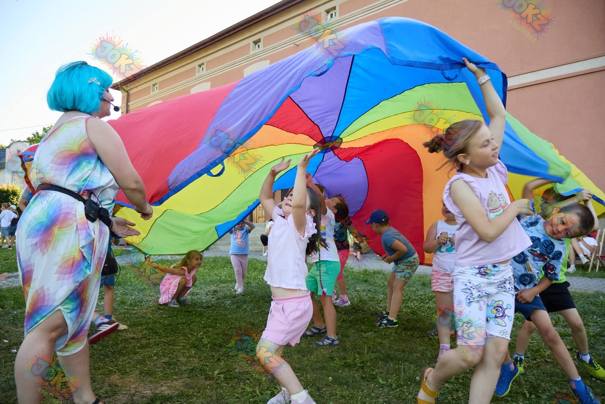 Animator z niebieskimi włosami i grupa dzieci bawią się kolorową chustą animacyjną na trawie. Zabawa na świeżym powietrzu, uśmiechy, energia i radość.