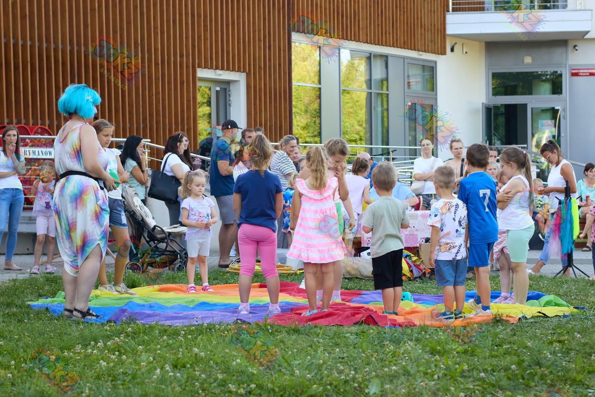 Animator z niebieskimi włosami prowadzi gry dla dzieci na kolorowej macie w Krosno. Rodzice obserwują zabawę na trawie przed budynkiem.