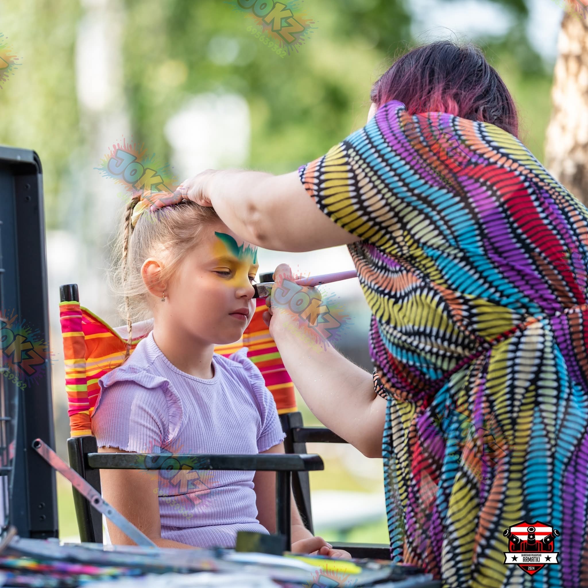 Animator maluje twarz dziewczynce w plenerze. Dziecko siedzi na krześle reżyserskim, a artystka w kolorowej sukience nakłada farby pędzlem. Widoczne farby i akcesoria.