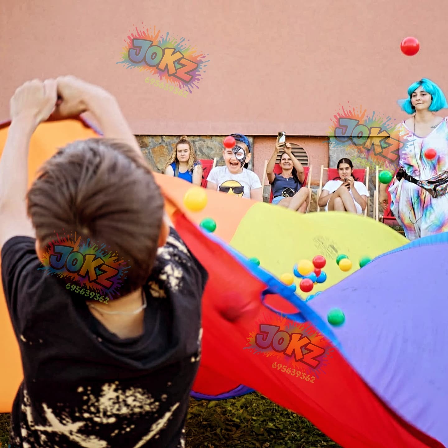 Animator prowadzi zabawę z kolorową chustą animacyjną i piłeczkami. Dzieci siedzą na leżakach w tle. Widoczne logo firmy JOKZ.