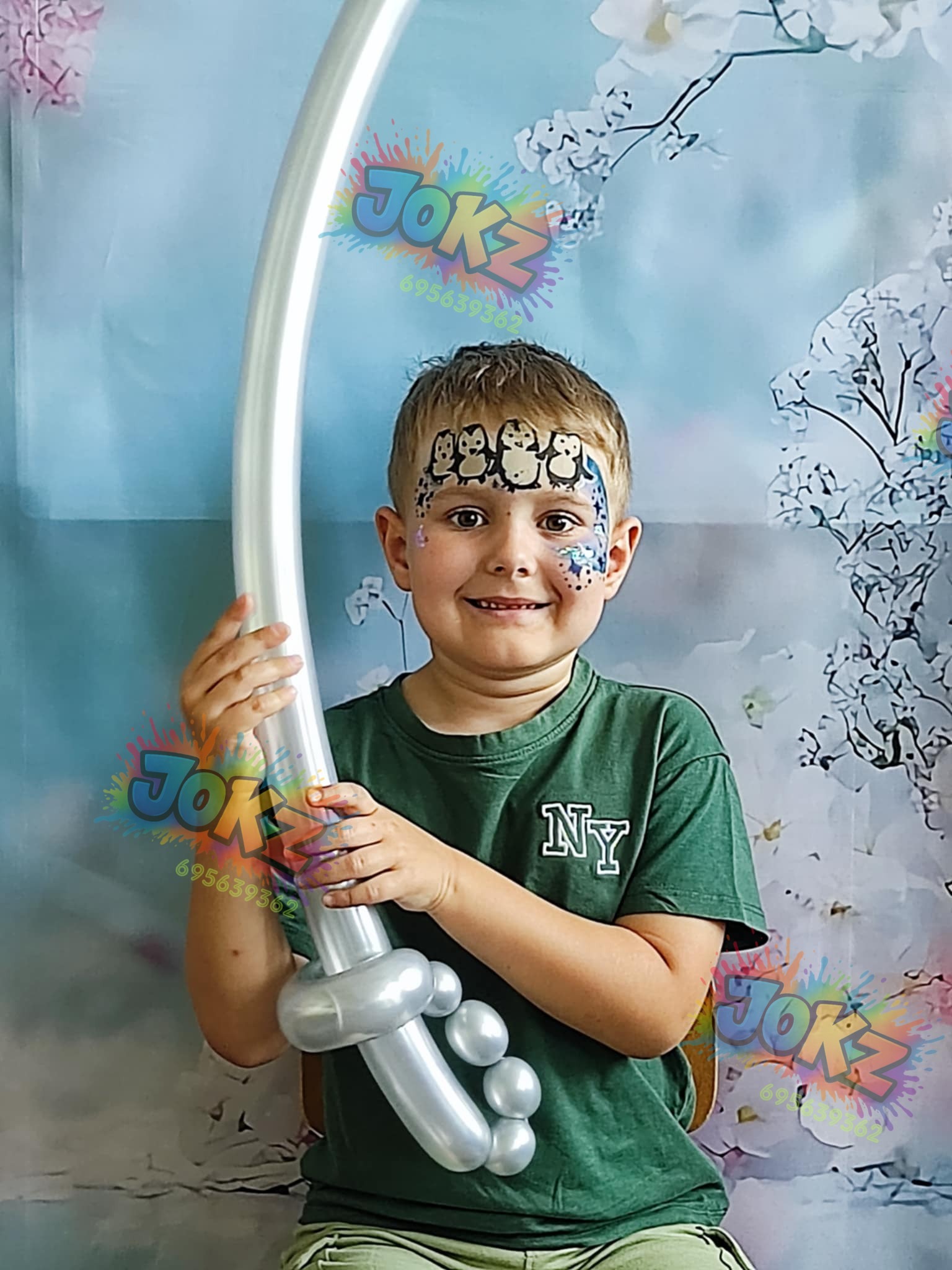 Uśmiechnięty chłopiec z pomalowaną twarzą (pingwiny) trzyma srebrny balon w kształcie miecza na tle dekoracji kwiatowej. Widoczny znak wodny.