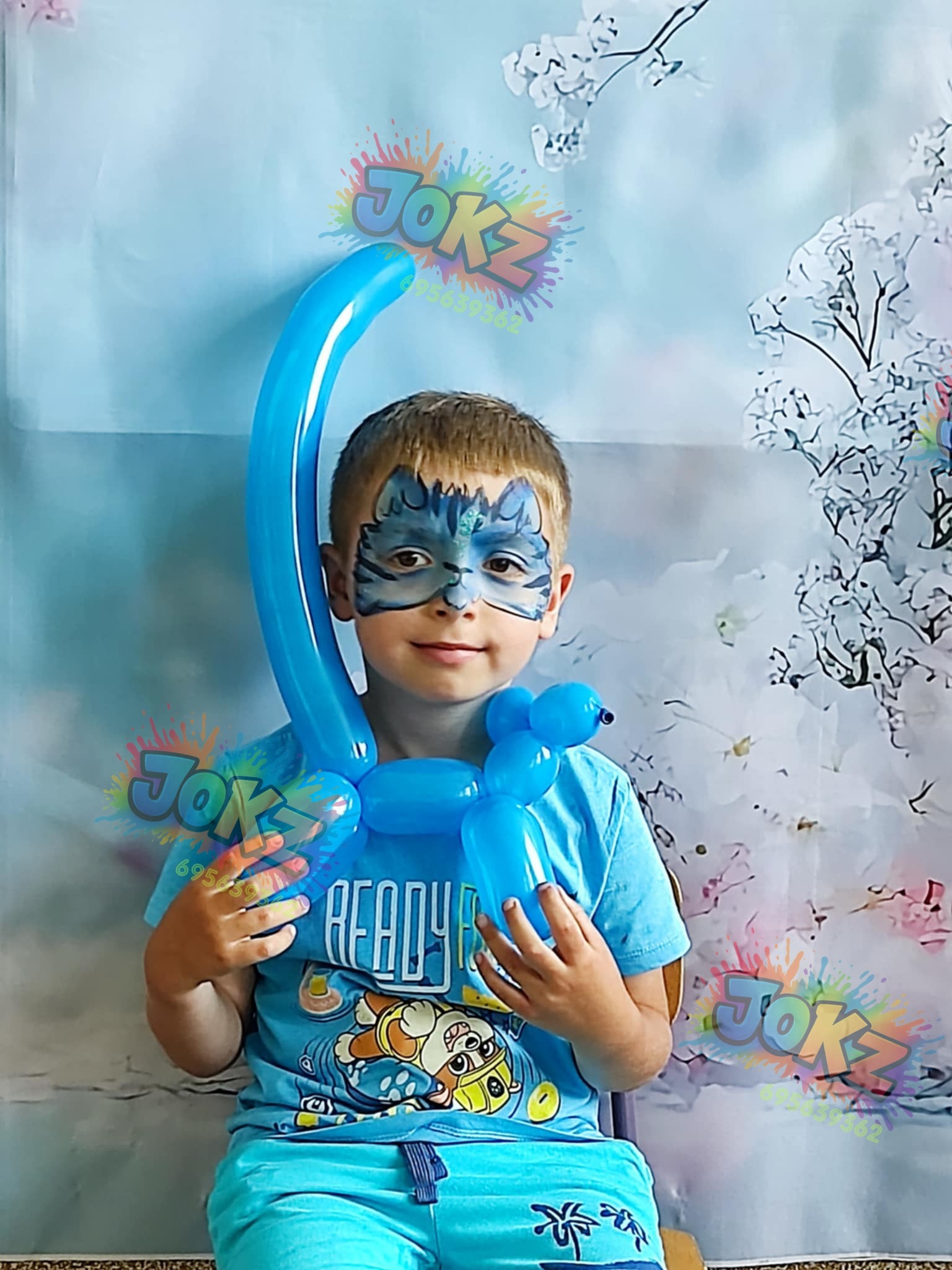 Uśmiechnięty chłopiec z pomalowaną twarzą w motywy zwierzęce, trzymający niebieskiego balonowego pieska i długi balon. Tło w kwiaty.