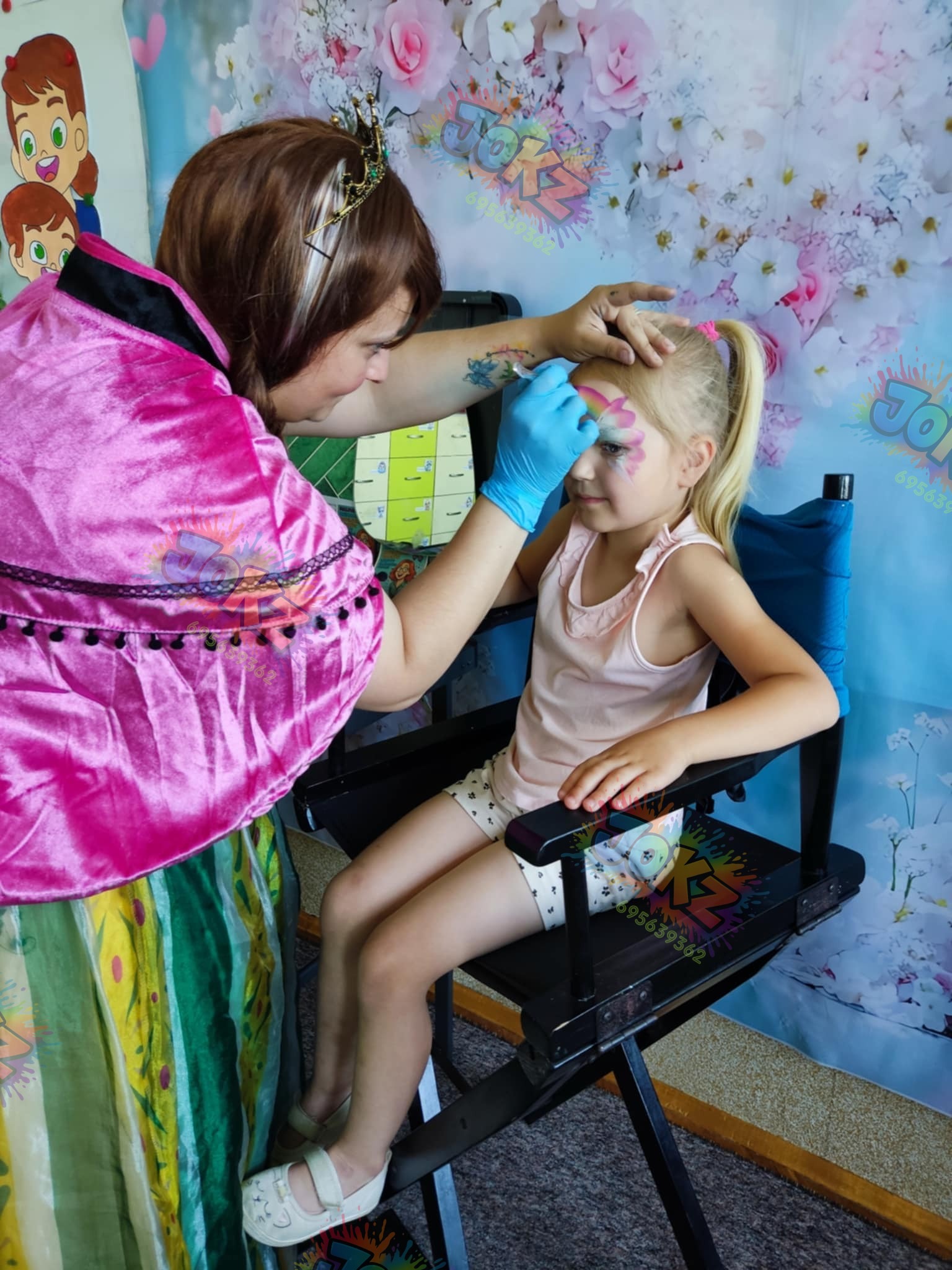 Animator maluje twarz dziewczynce w fotelu reżyserskim. Tło w kwiaty, kostium animatora w odcieniach różu i zieleni. Na czole dziecka tęcza i kwiat.