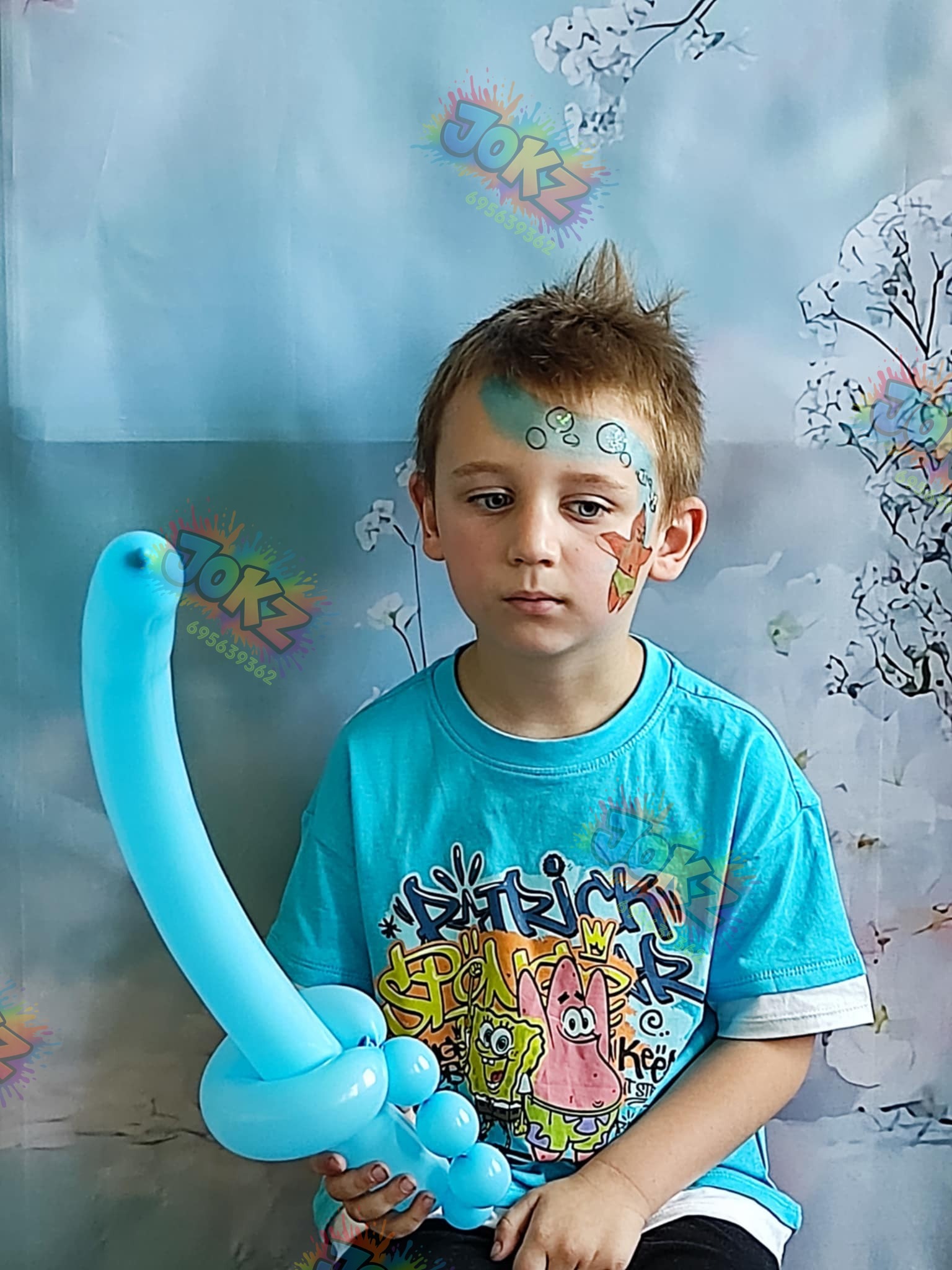 Chłopiec z malunkiem na twarzy (motywy morskie) trzyma niebieskiego balona w kształcie łabędzia. Tło w odcieniach błękitu z motywem kwitnących gałęzi.