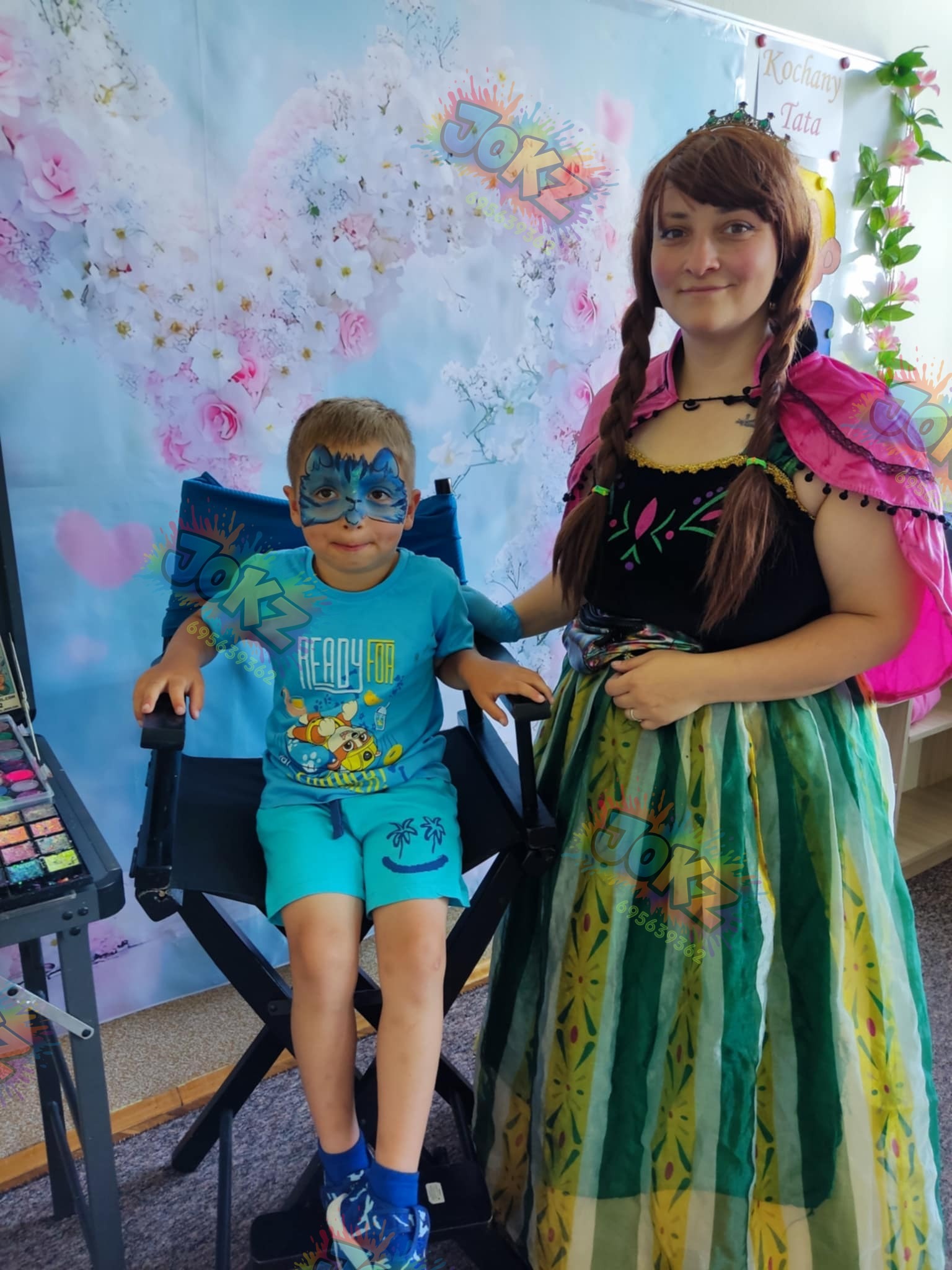 Animator w stroju Anny z Krainy Lodu maluje twarz chłopcu w niebieskiej koszulce, siedzącemu na krześle reżyserskim. W tle kwiatowa dekoracja.