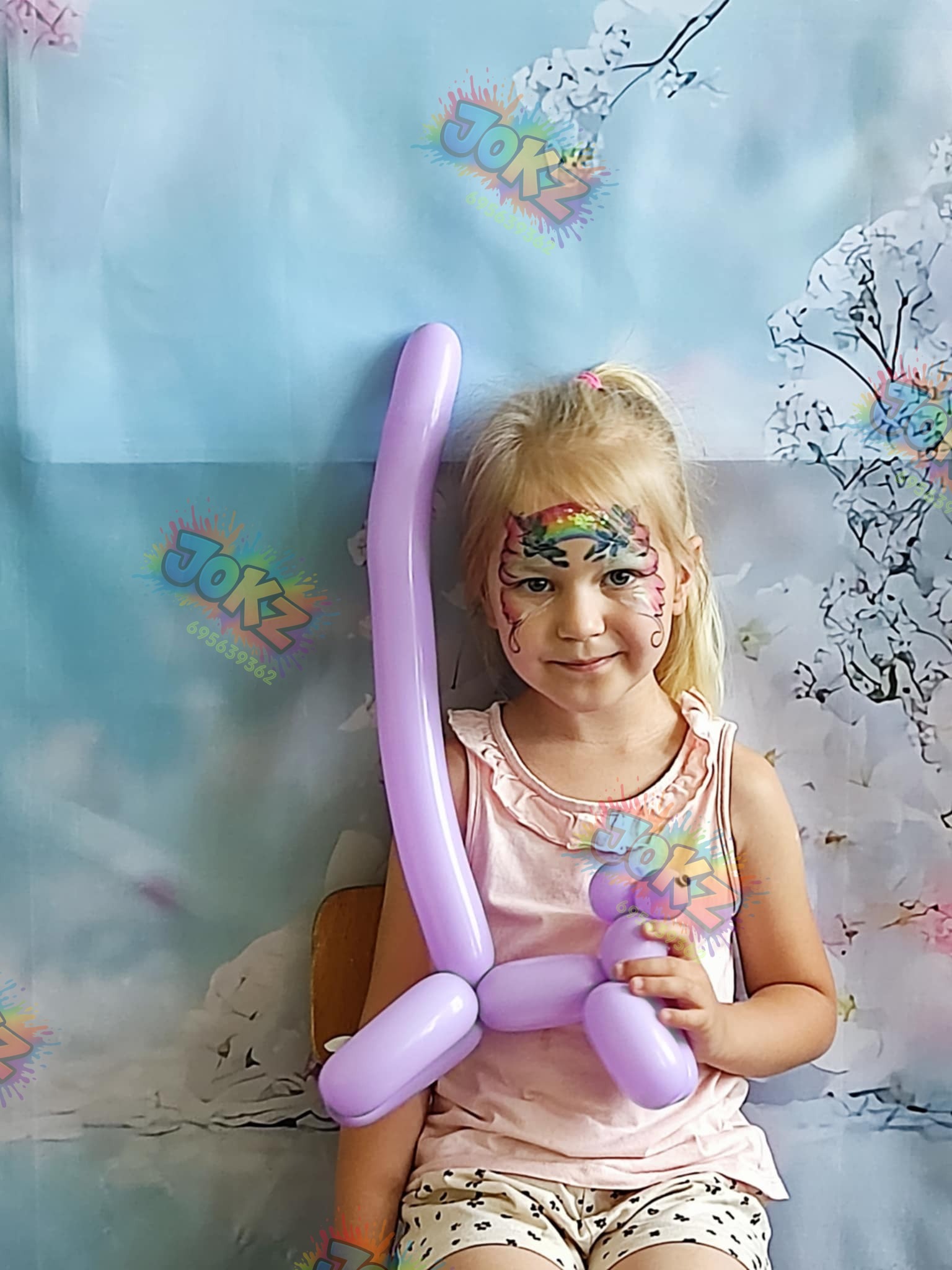 Uśmiechnięta blondynka z tęczowym malunkiem na czole trzyma fioletowego balonowego zwierzaka na tle dekoracji z motywem kwiatowym.