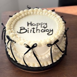 Kruche inspiracje - Tort urodzinowy z kremową dekoracją i czarnymi wstążkami, umieszczony na drewnianym stole w naturalnym świetle. Napis 'Happy Birthday' wykonany czarną czcionką.