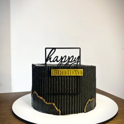 Kruche inspiracje - Czarny tort urodzinowy z teksturą, zdobiony złotem i napisem 'Happy Birthday', na białym podeście i drewnianym stole. Minimalistyczny design.