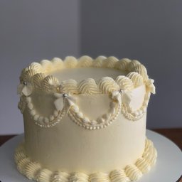 Kruche inspiracje - Elegancki tort w stylu vintage z kremową dekoracją, zdobiony srebrnymi koralikami i rozetkami, na drewnianym podeście. Subtelne oświetlenie podkreśla detale.