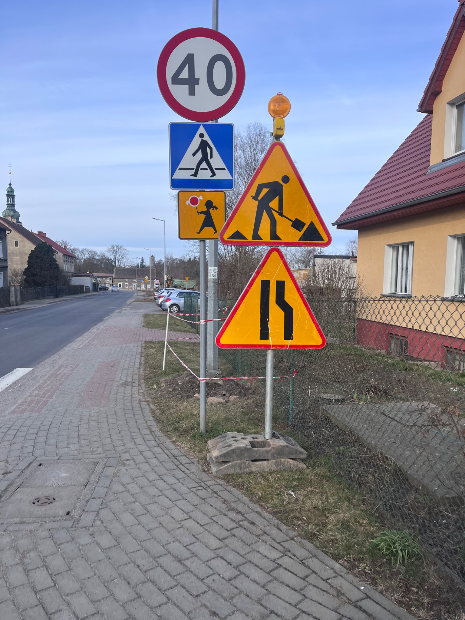 Znak drogowy ostrzegający o robotach i zwężeniu jezdni, ustawiony przy chodniku z kostki. W tle budynki mieszkalne i błękitne niebo.